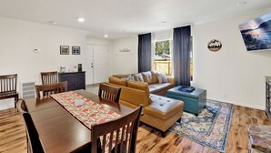 Una sala de estar acogedora con un sofá, un reposapiés y una mesa de comedor con sillas. Hay una ventana con cortinas, una obra de arte colgada en la pared y un letrero con una montaña y árboles.