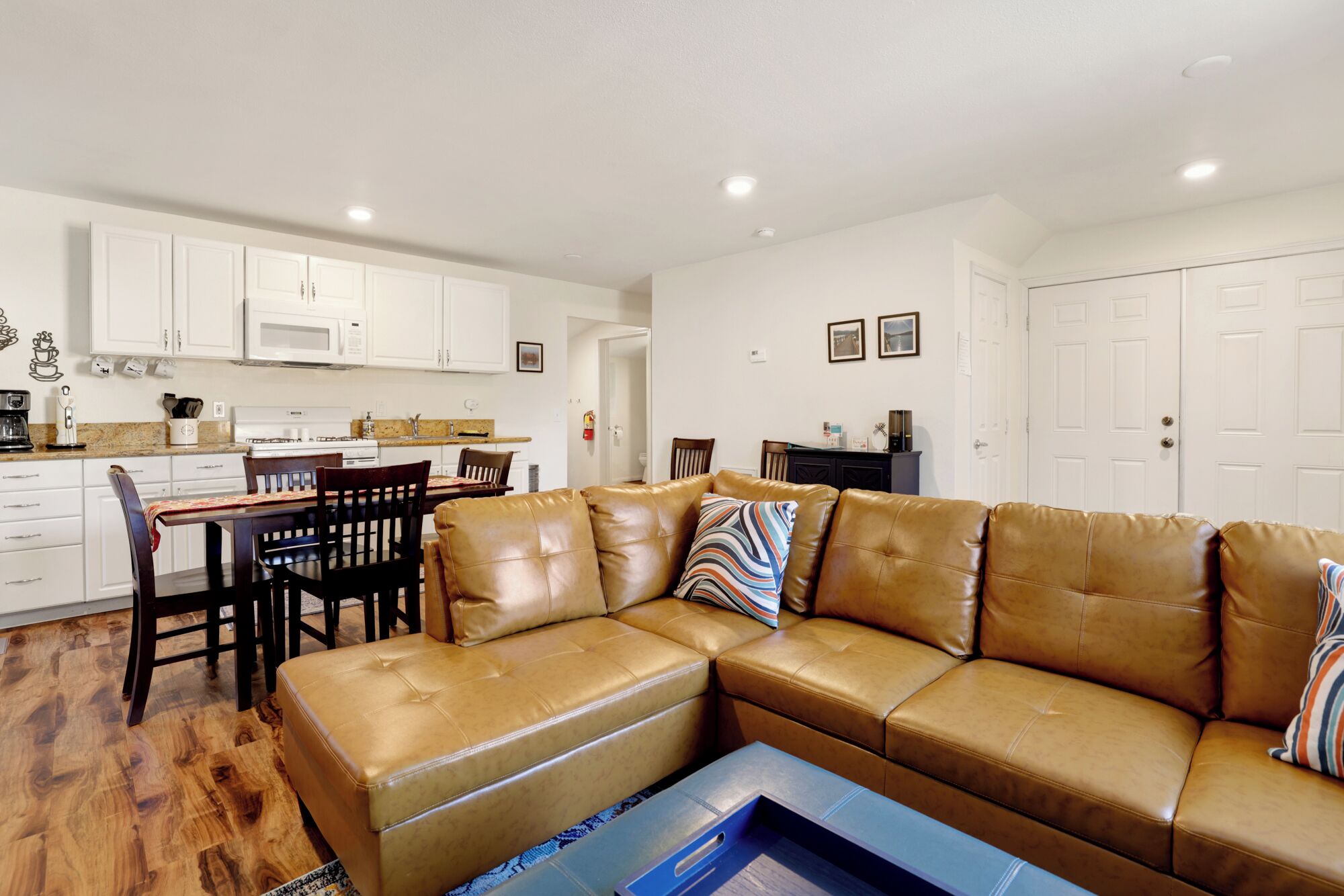 A living room with a brown leather sectional sofa, a dining table with chairs, a kitchen area with white cabinets, and a door leading to another room.