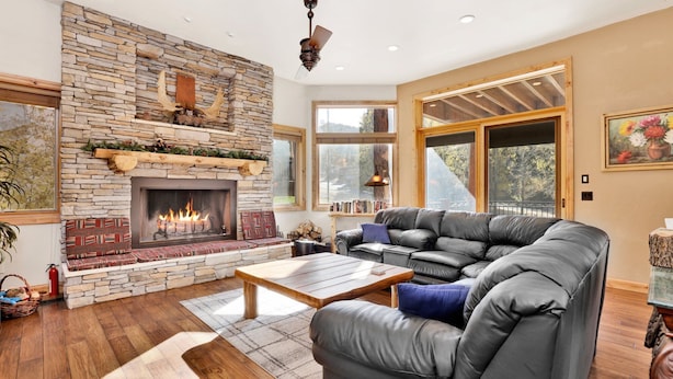 A cozy living room with a stone fireplace, leather sofa, and large windows offering a view of the outdoors.