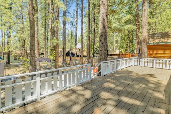 A wooden deck with a white railing, surrounded by tall pine trees.