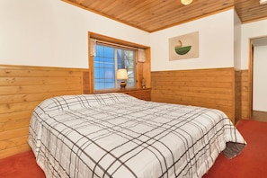 A bedroom with a wooden headboard, a window with blinds, a bedside lamp, and a wall-mounted artwork.