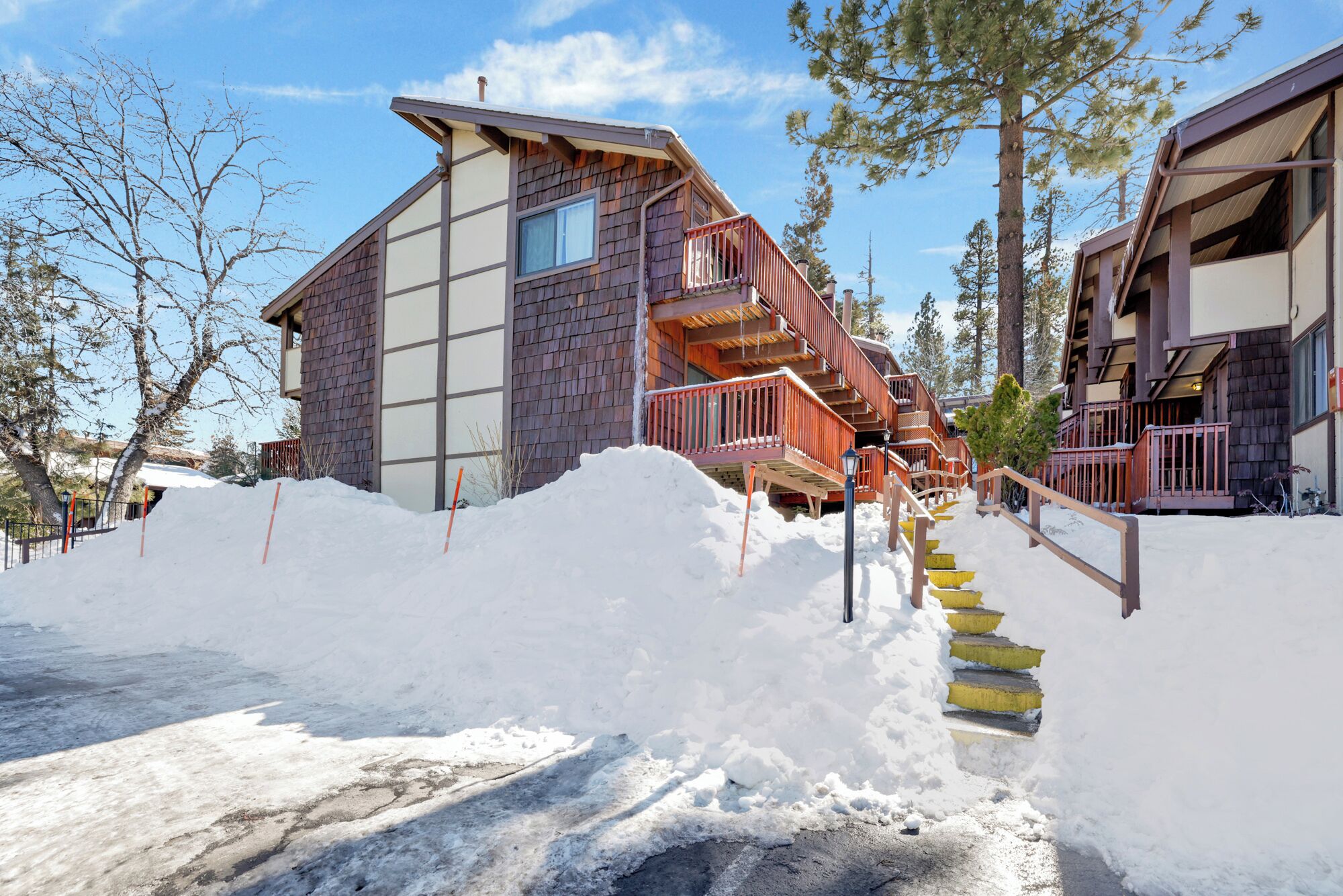 A wooden cabin with a staircase leading to a deck, surrounded by snow and trees.