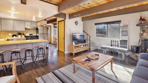 A spacious living area with a wooden ceiling, a kitchen with modern appliances, a large window with a view, and a fireplace. - Pinewood INN #2093 (Big Bear City)