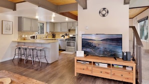 A modern kitchen with a central island, bar stools, and a large flat-screen TV on a wooden stand. - Pinewood INN #2093 (Big Bear City)