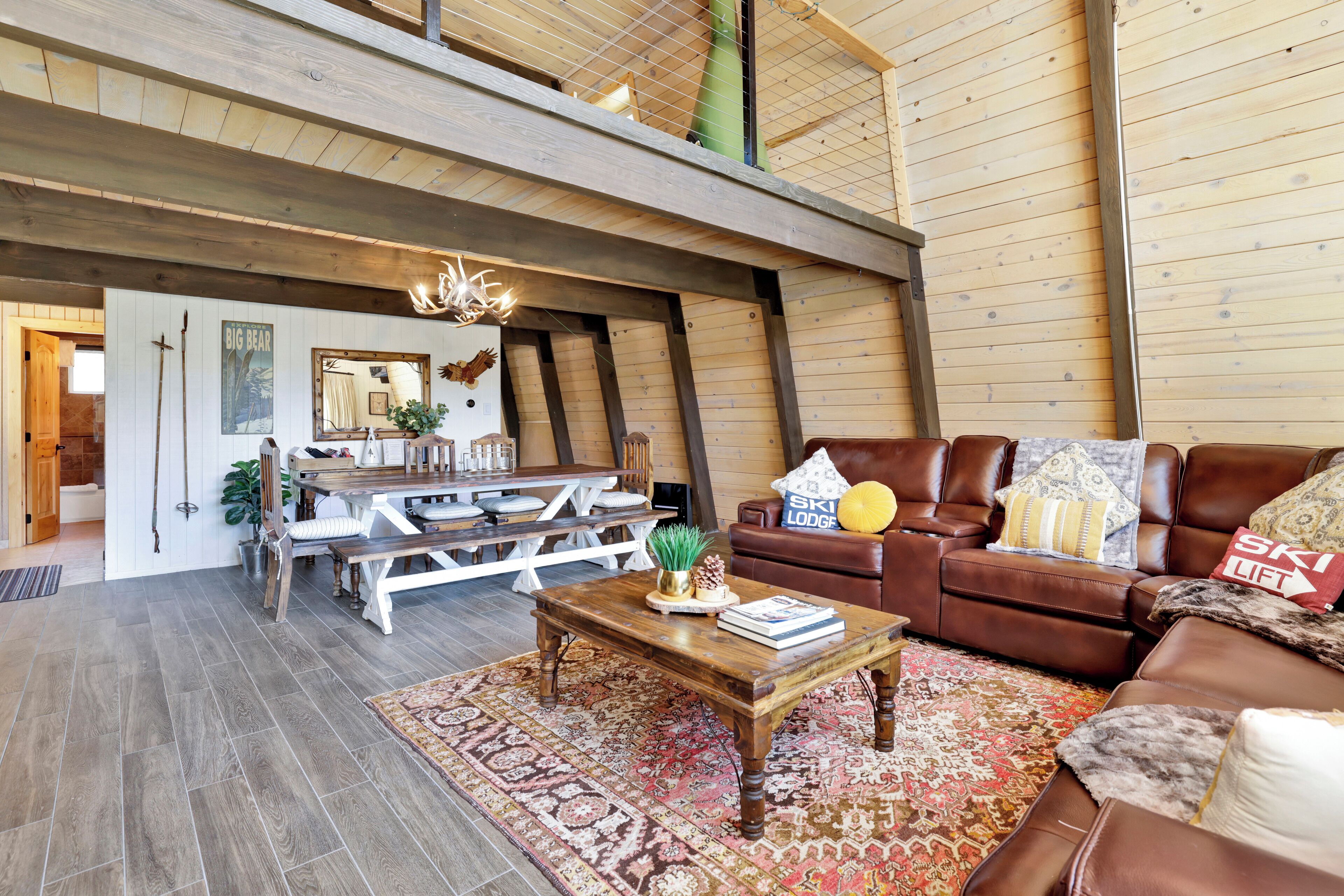 A spacious loft-style living room with a high ceiling, wooden beams, a brown leather sofa, a wooden coffee table, and a patterned rug.