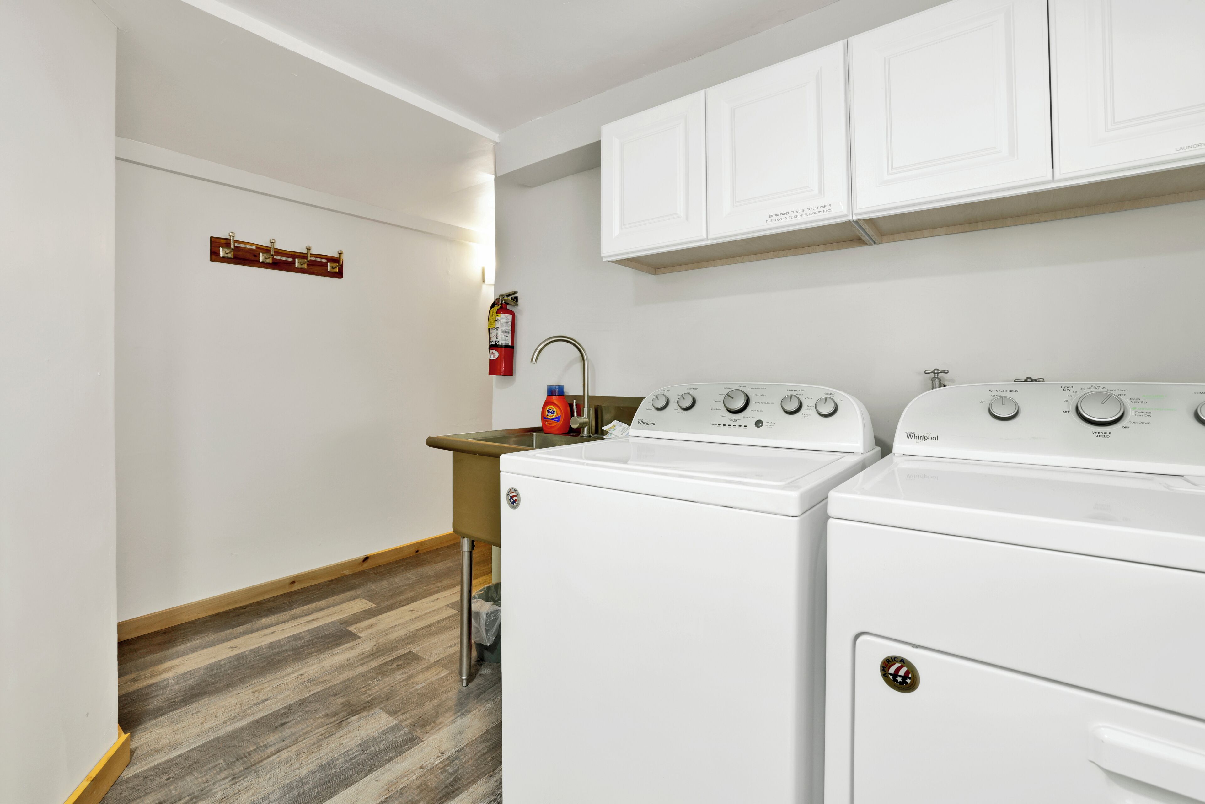 A laundry room with two white Whirlpool washers and dryers, a wooden floor, and white cabinets above the appliances.
