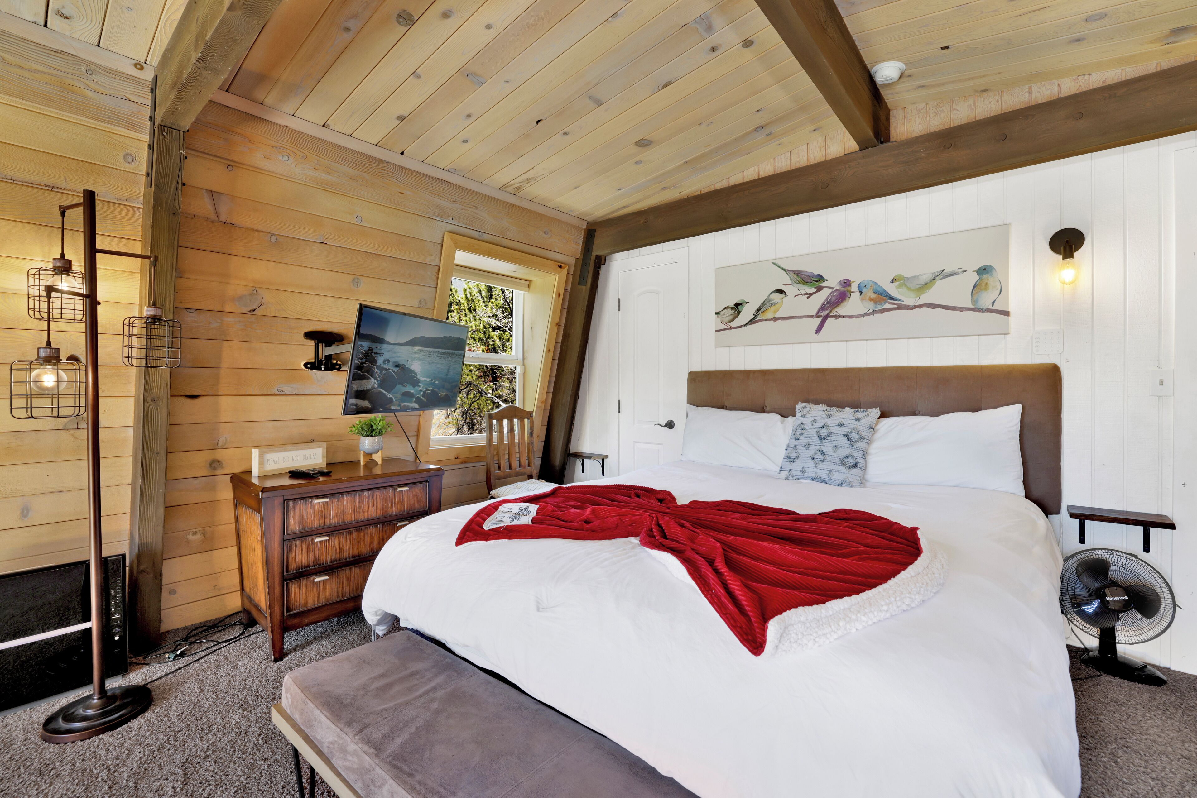 Een slaapkamer met een houten plafond, een bed met witte beddengoed en een rode deken, een houten dressoir, een bureau met een lamp, en een schilderij van vogels aan de muur.
