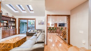 A spacious living area with a leather sofa, wooden coffee table, and a bookshelf. There are skylights on the ceiling, a dining area with chairs, and a TV mounted on the wall. - Bears Palace Chalet #2008 (Big Bear Lake)