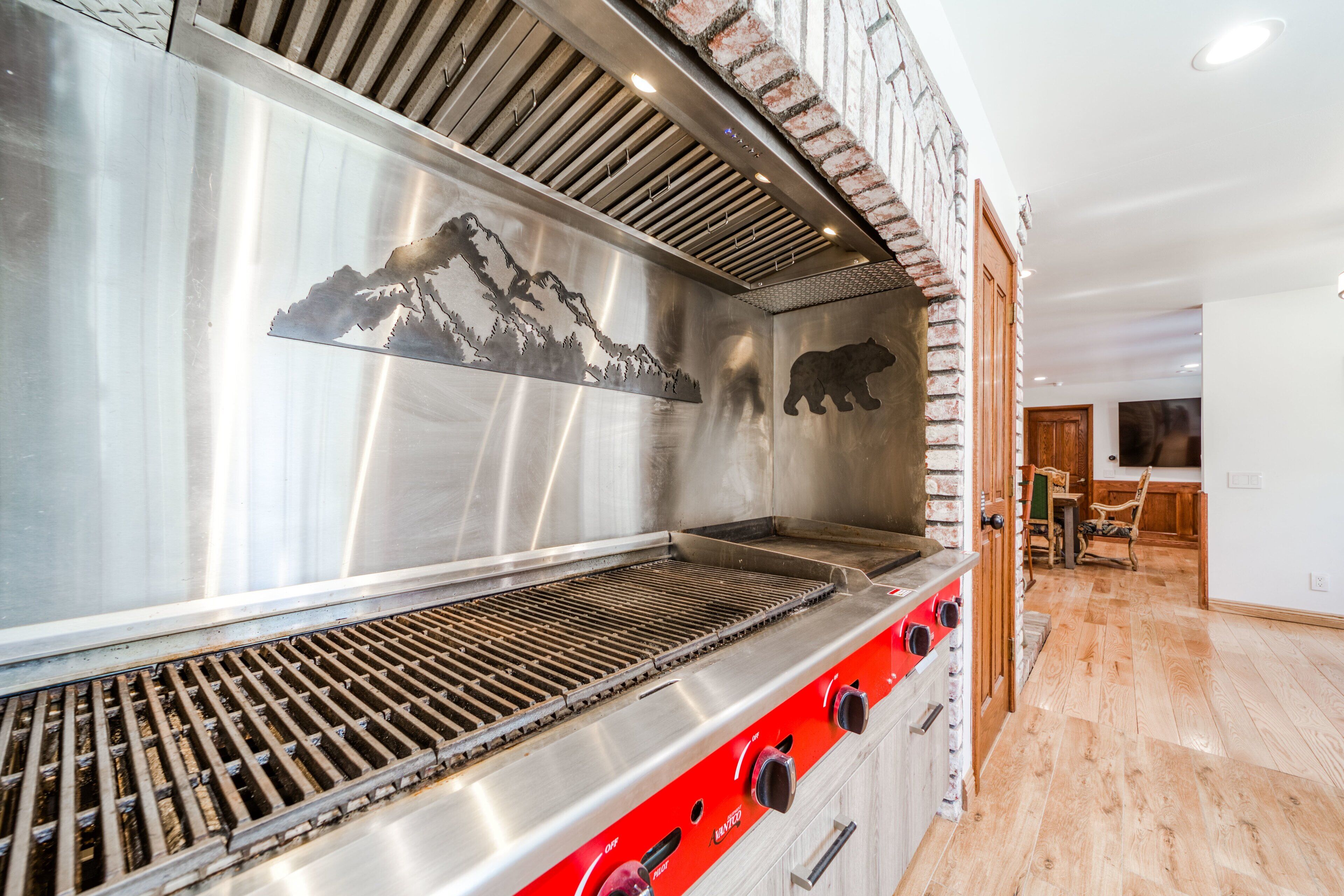 Una parrilla de acero inoxidable con silueta de oso y diseño de montaña, suelo de madera y pared de ladrillo.