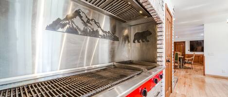 Una parrilla de acero inoxidable con silueta de oso y diseño de montaña, suelo de madera y pared de ladrillo.