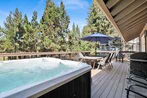 A hot tub on a wooden deck with a view of tall trees, outdoor furniture, and a covered area. - ALL About FUN #1149 (Big Bear Lake)