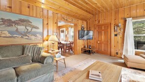 Een gezellig interieur van een chalet, met houten inrichting, een bank, een bijzettafeltje, een televisie en een schilderij van een boom.