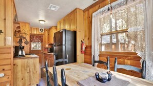 Une cabane en bois avec une table à manger, des chaises, un réfrigérateur et une fenêtre ornée de rideaux en dentelle.