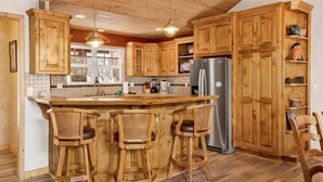 A kitchen with wooden cabinets, a stainless steel refrigerator, and a bar area with stools. - ONE Hundred Pines Ranch #2005 (Big Bear City)