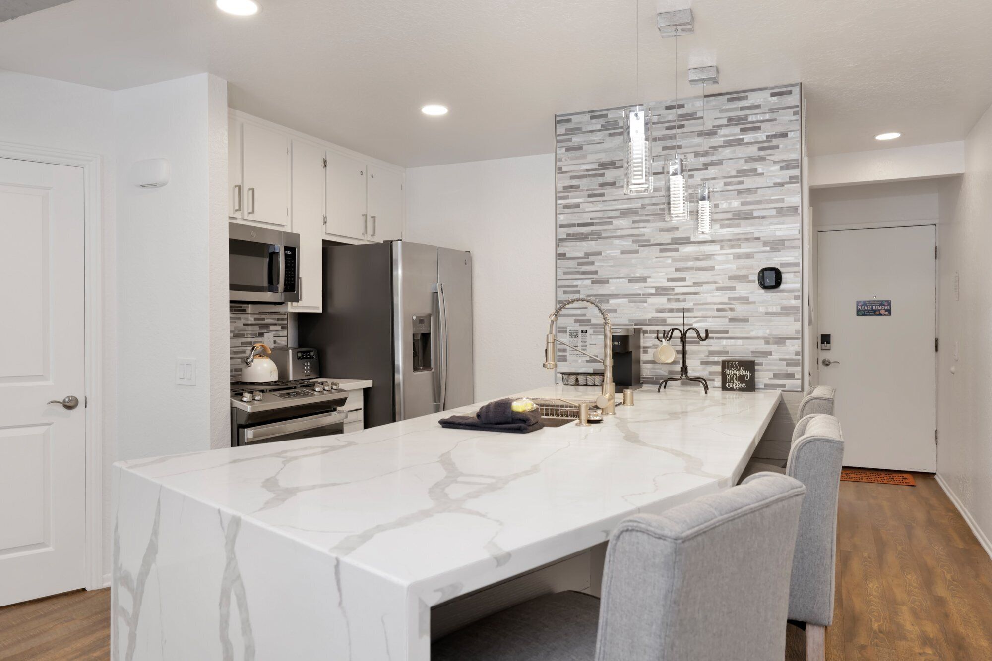 A modern kitchen with white cabinetry, a marble island, stainless steel appliances, and a gray backsplash.