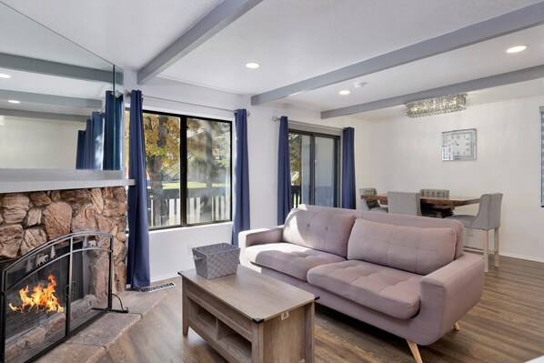 A modern living room with a stone fireplace, a beige sofa, and a dining area with a wooden table and chairs. - Blue JAY Getaway #2082 (Big Bear Lake)
