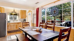 A cozy kitchen with wooden cabinets, a dining table with papers, and a large window with a view of trees. - Montana Cabin #1938 (Big Bear City)