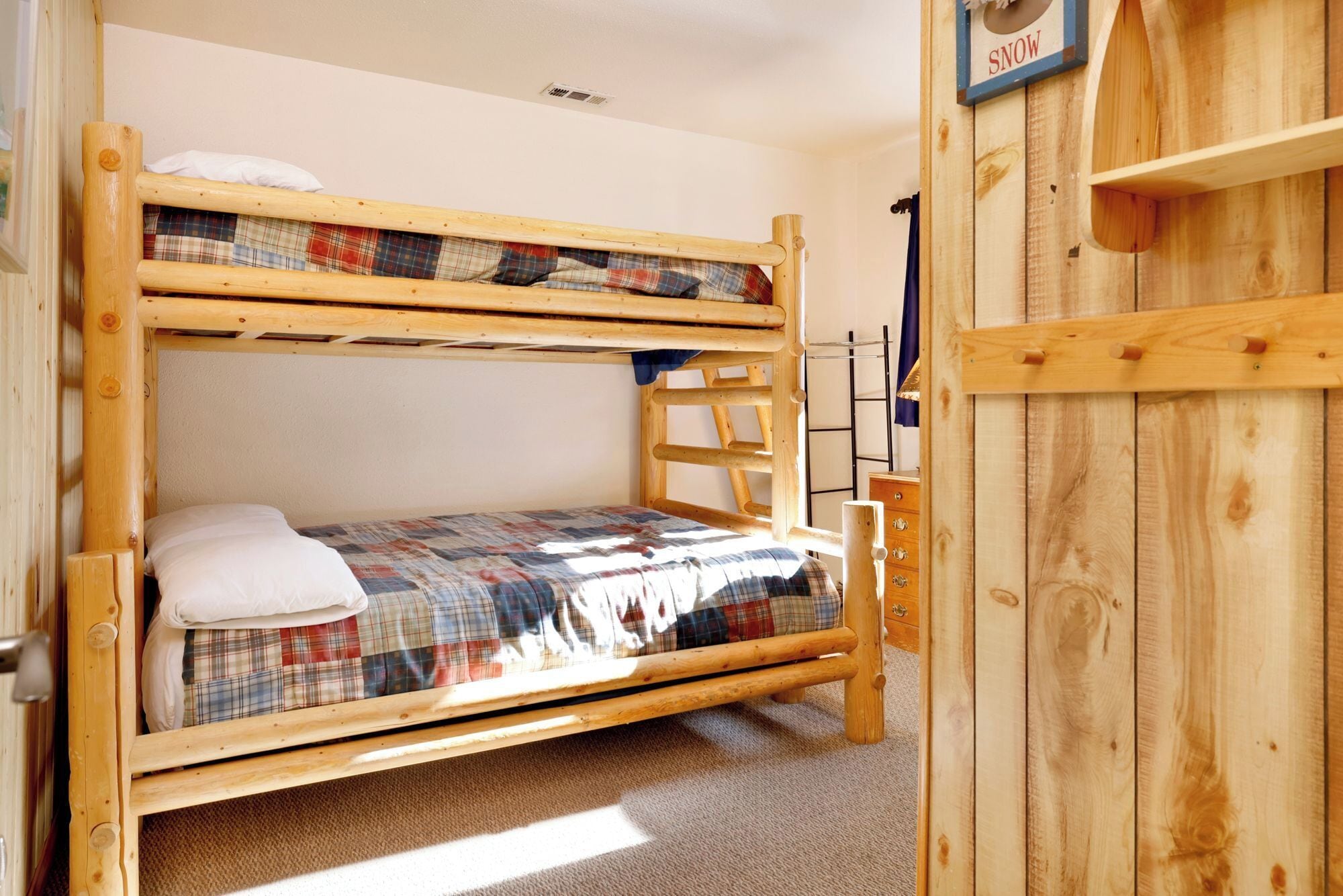Un lit superposé avec une couette à carreaux, une échelle en bois et une petite étagère en bois sur laquelle est accrochée une pancarte indiquant « NEIGE ».