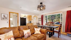 A cozy living room with a brown leather sofa, a dining table with chairs, a kitchen in the background, and a large window with curtains. - Montana Cabin #1938 (Big Bear City)