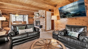 L’intérieur d’une cabane en rondins, avec une cheminée, des canapés en cuir, un coin repas et une grande fenêtre.