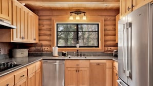 Une cuisine rustique aménagée dans une cabane en rondins, avec des meubles en bois, un réfrigérateur en acier inoxydable, un lave-vaisselle et une fenêtre donnant sur la forêt.