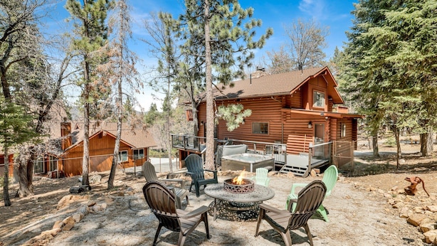 Une cabane en rondins avec une terrasse, entourée d’arbres. On y trouve un foyer avec des chaises disposées devant.