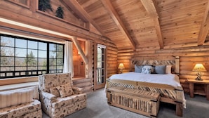 Une chambre dans un chalet en rondins, avec un grand lit, un fauteuil à motifs animaliers, une fenêtre donnant sur les arbres et une lampe sur la table de chevet.