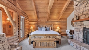 Une chambre à coucher aménagée dans un chalet en rondins, avec un lit, des lampes de chevet, une cheminée en pierre et un banc en bois rustique.