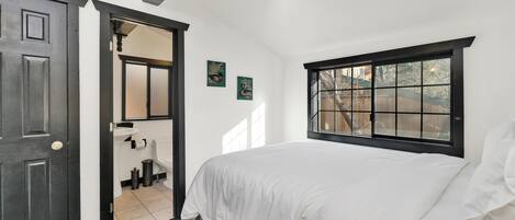 A bedroom with a bed, a window, a door, and a bathroom visible through an open door.