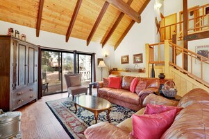 A spacious living room with a high wooden ceiling, a leather sofa, a glass door leading to an outdoor area, and a staircase with wooden railings.