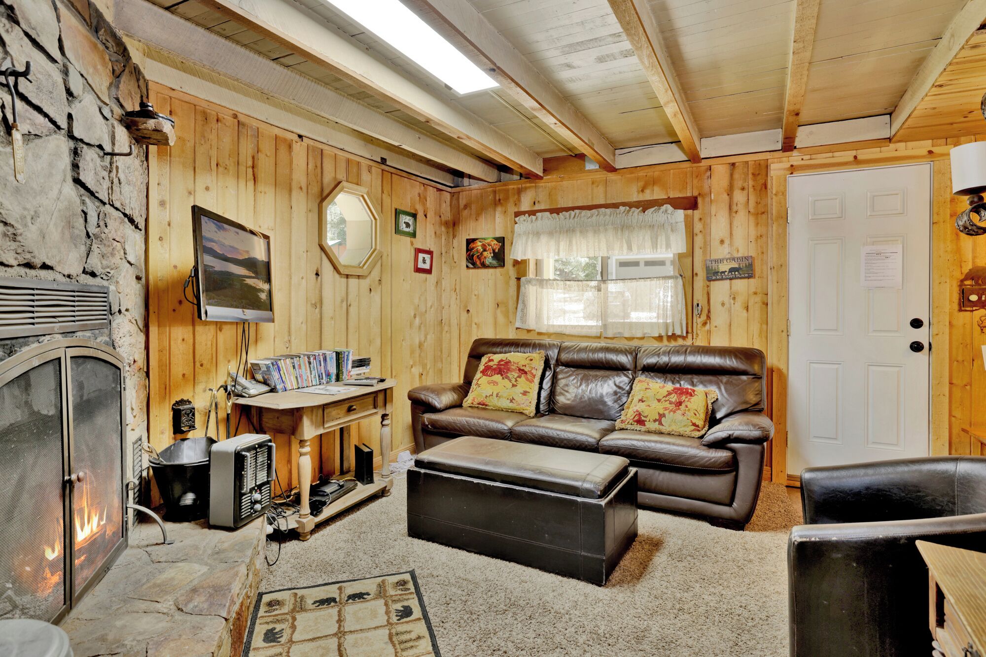 A cozy cabin interior with a stone fireplace, wooden walls, a leather sofa, and a small table with books and a radio.