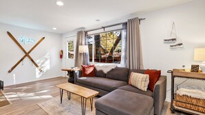 A modern living room with a grey sectional sofa, a wooden coffee table, and a view of trees through large windows. - Apres Chalet #2364 (Sugarloaf)