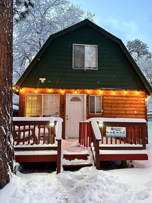 Une cabane en rondins avec un porche enneigé et une pancarte indiquant une capacité maximale de 6 personnes et de 2 voitures.