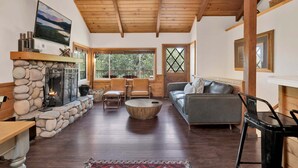 A cozy living room with a stone fireplace, wooden beams, a sofa, and a large window. - Black OAK Cabin #1977 (Big Bear City)