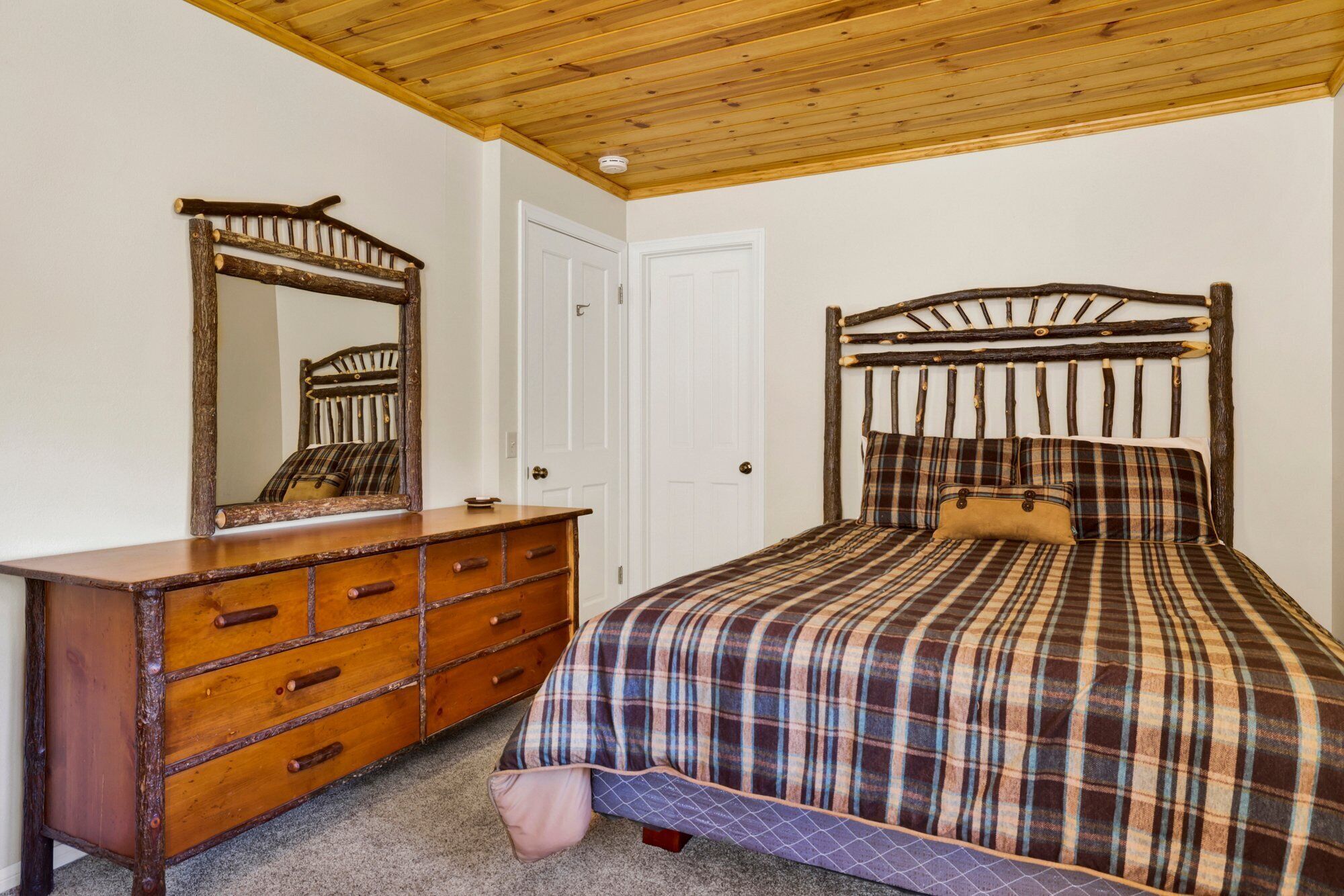 A bedroom with a wooden headboard, a wooden dresser with drawers, a plaid bedspread, and a mirror on the dresser.
