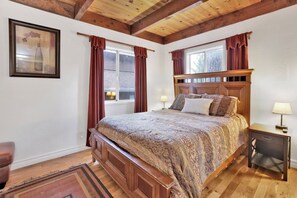 A bedroom with a wooden bed, two bedside tables with lamps, a window with curtains, and a framed picture on the wall. - The Cozy Cottage#2384 (Big Bear City)