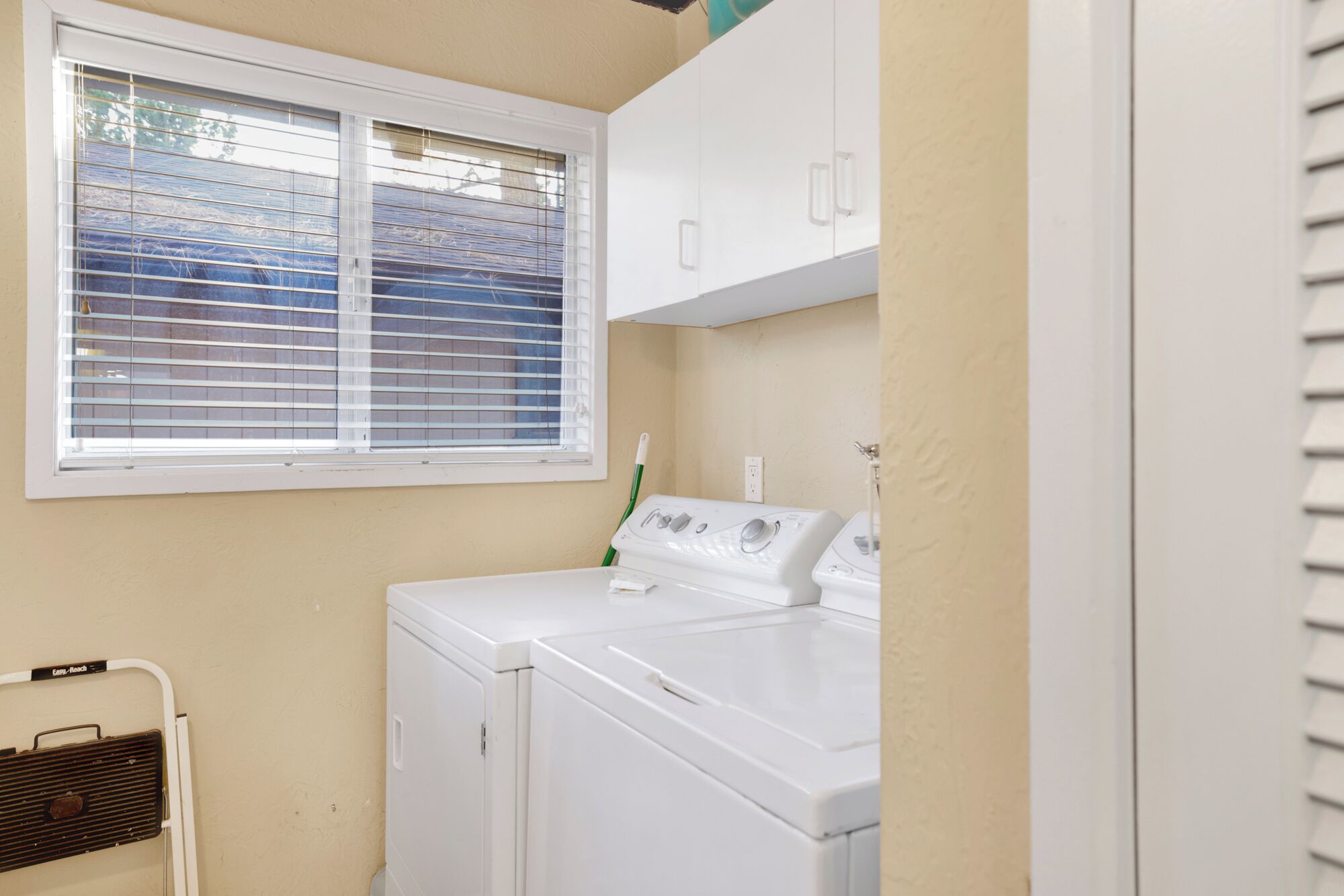 A laundry room with a front-loading washer and dryer, a window with blinds, and a white cabinet above the appliances.
