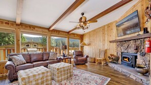 Ein gemütliches Blockhaus-Interieur mit Kamin, Holzwänden, einer Ledersofa und karierten Hocker, mit einem großen Fenster mit Blick auf einen bewaldeten Bereich.