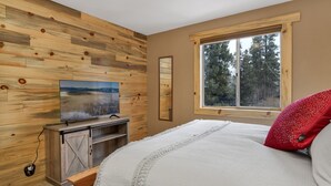 A bedroom with a wooden wall, a large window with a forest view, a TV on a wooden stand, and a bed with a white and red pillow. - Emma's Cottage#2267 (Big Bear City)