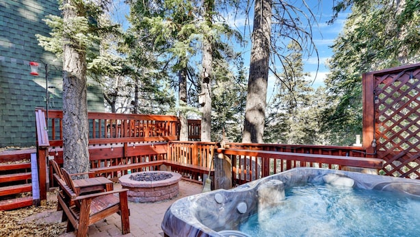 A hot tub surrounded by wooden deck seating, a fire pit, and tall trees in a backyard setting.