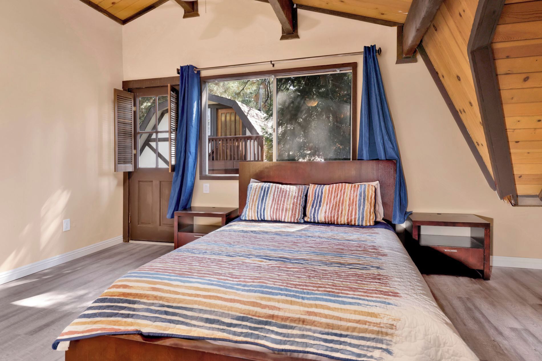 A bedroom with a wooden bed, striped bedding, and a view of a wooden deck and trees through large windows.