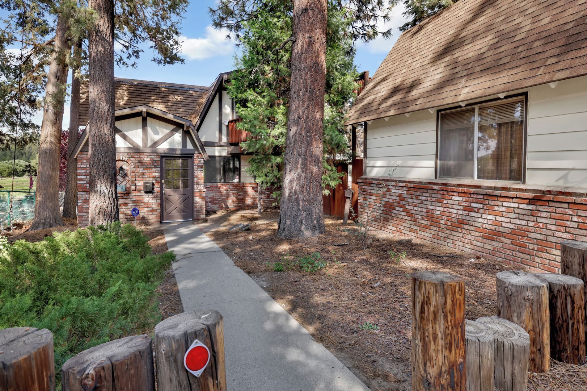 A rustic cabin with a brick exterior and a wooden roof, surrounded by tall trees and a gravel path.