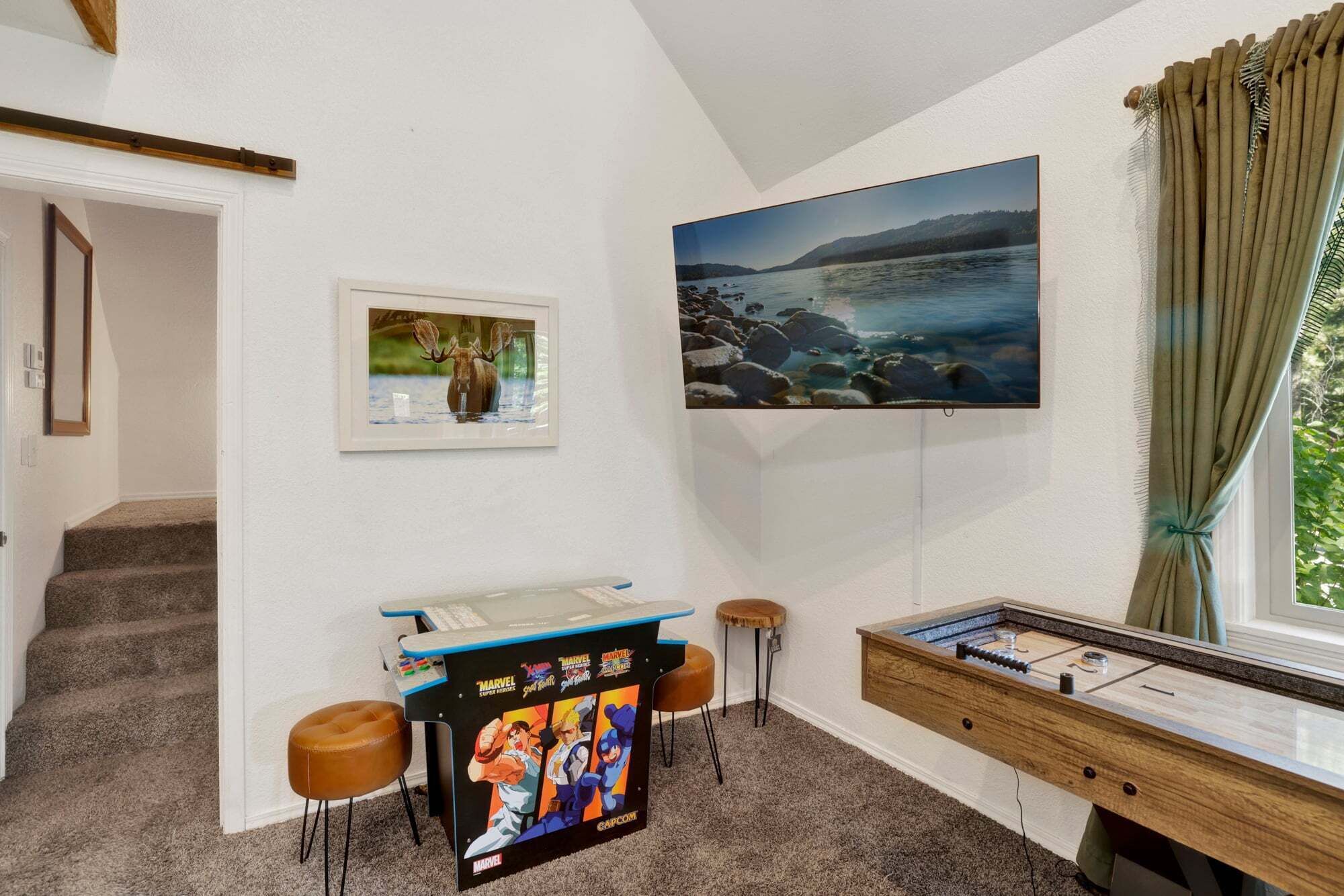A room with a desk, a mounted TV, a framed picture, a window with curtains, and a staircase.