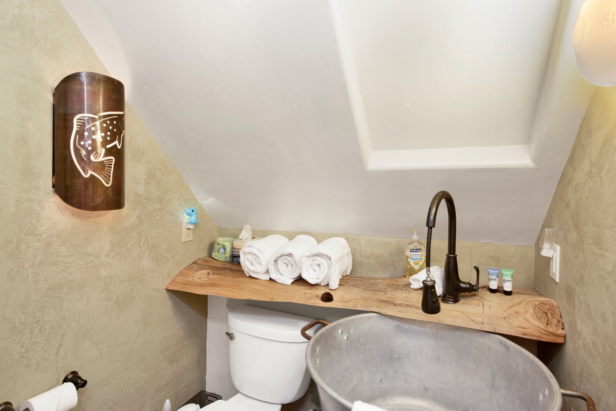 A bathroom with a wooden shelf holding towels and toiletries, a toilet, and a metal basin.