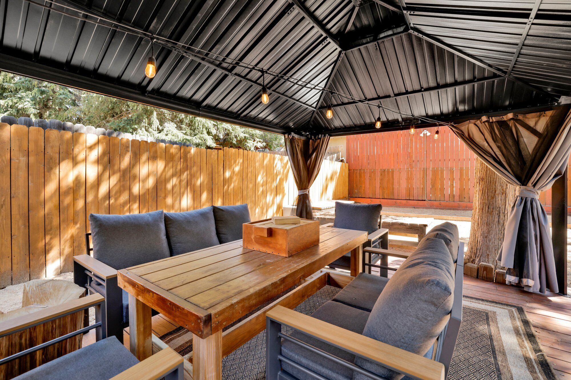 Eine Terrasse im Freien mit Holztisch und -stühlen, einer gepolsterten Bank und einem überdachten Bereich mit Metallveranda.