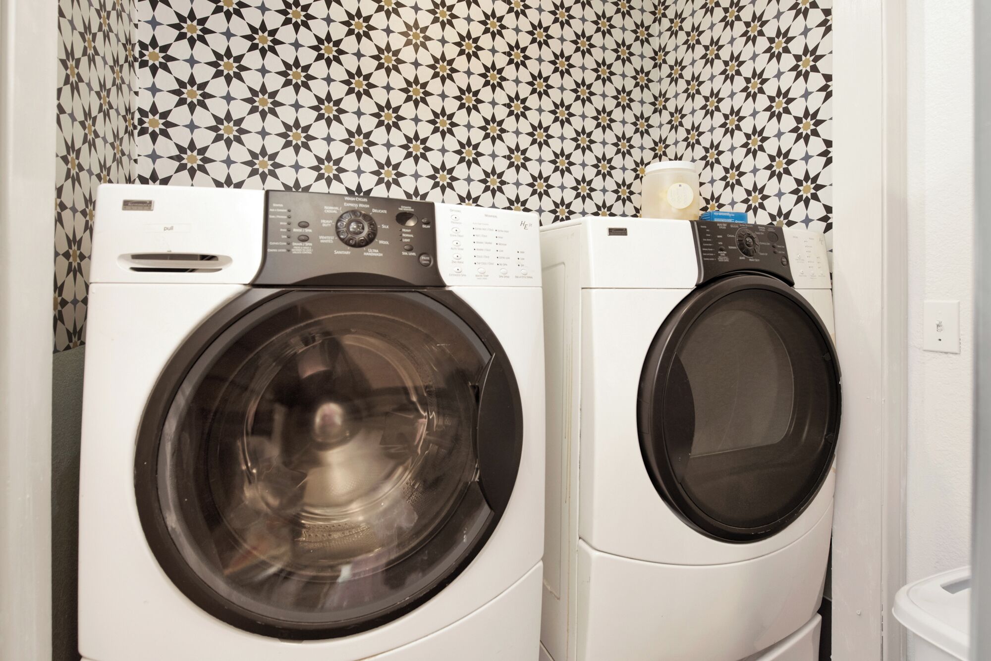 Two front-loading washing machines in a laundry area with patterned wallpaper.