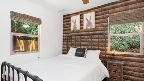 A bedroom with a wooden log wall, a bed with white bedding, two windows with blinds, and a nightstand with a lamp. - Maltby Modern LOG Cabin #2307 (Big Bear City)