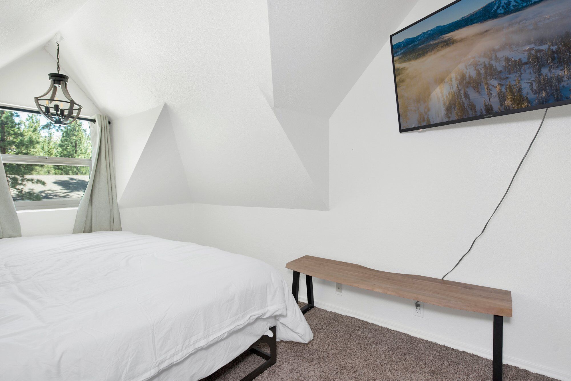 Un dormitorio con una cama, un lámpara colgante, un televisor montado y un banco.