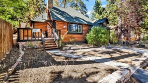 A rustic log cabin with a stone pathway leading to the entrance, surrounded by trees and a fenced garden area. - Maltby Modern LOG Cabin #2307 (Big Bear City)