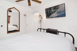 A bedroom with a bed, a large mirror, a ceiling fan, a TV mounted on the wall, and a framed picture of a snowy landscape.